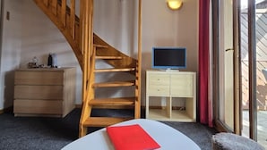 A room with a wooden staircase, a flat-screen TV, a small white table with a red book, a fluffy stool, and a sliding glass door to the outside.