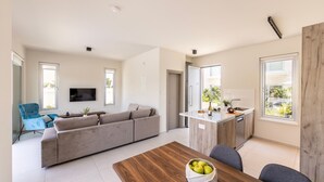 A modern living room with a sofa, a dining table with a bowl of fruit, a kitchen area with a sink and countertop, and large windows. - Protaras Mythical Seas (Paralimni)