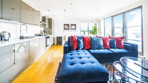 A modern kitchen with a dining area, a blue sofa with red and blue cushions, and a large window offering a view of the city. - Modern 2BR in Surrey | Pet-Friendly, WiFi, Parking (Surrey)