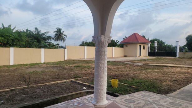 A courtyard with a decorative pillar, a small building with a red roof, and a beige wall. - Assembly Hotel - Esiama  (Esiama)