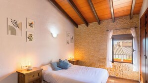 A bedroom with a stone wall, a wooden ceiling, a bed with white bedding, a nightstand with a lamp, and a window with a view of a tiled roof.