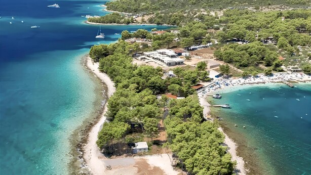 Ett flygbildsbild av ett kustområde med klart turkosfärgat vatten, en sandstrand och en liten ö med tät vegetation och byggnader.