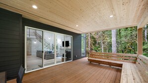 Une terrasse en bois avec un banc et une porte vitrée donnant sur un espace de vie comprenant un canapé et un téléviseur.