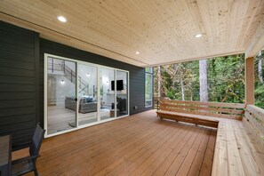 A wooden deck with a bench and a glass door leading to a living area with a sofa and a TV. - Sandy River Group Getaway 4 (Mount Hood Village)