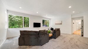 Un salon spacieux avec un grand canapé d’angle, un téléviseur à écran plat fixé au mur et une grande fenêtre donnant sur un paysage verdoyant.