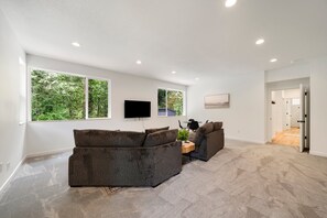 A spacious living room with a large sectional sofa, a flat-screen TV mounted on the wall, and a large window offering a view of greenery. - Sandy River Group Getaway 4 (Mount Hood Village)