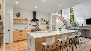 Une cuisine moderne dotée d’un îlot en marbre, de tabourets de bar et d’un escalier menant à un espace de vie comprenant un canapé et un téléviseur.