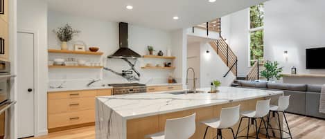 A modern kitchen with a marble island, bar stools, and a staircase leading to a living area with a sofa and a TV.