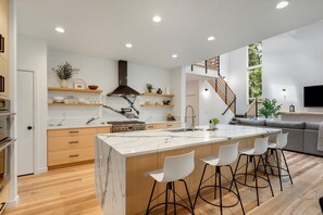 A modern kitchen with a marble island, bar stools, and a staircase leading to a living area with a sofa and a TV. - Sandy River Group Getaway 4 (Mount Hood Village)