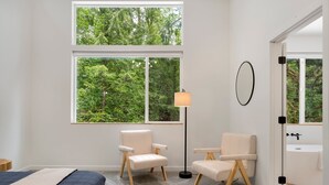 A bedroom with a large window, two chairs, a floor lamp, and a view of a forest. - Sandy River Group Getaway 4 (Mount Hood Village)