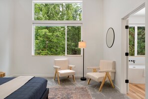 A bedroom with a large window, two chairs, a floor lamp, and a view of a forest. - Sandy River Group Getaway 4 (Mount Hood Village)