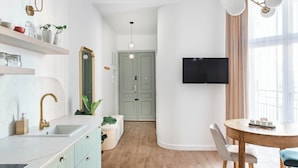Une cuisine moderne avec un plan de travail blanc, un robinet doré et des meubles en bois. On y trouve un coin repas avec une table ronde et des chaises, ainsi qu’un téléviseur fixé au mur.