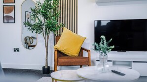 A modern living room with a brown leather chair, a yellow pillow, a round white coffee table, a large flat-screen TV, and a potted plant. - Elm Apartments (Salford)