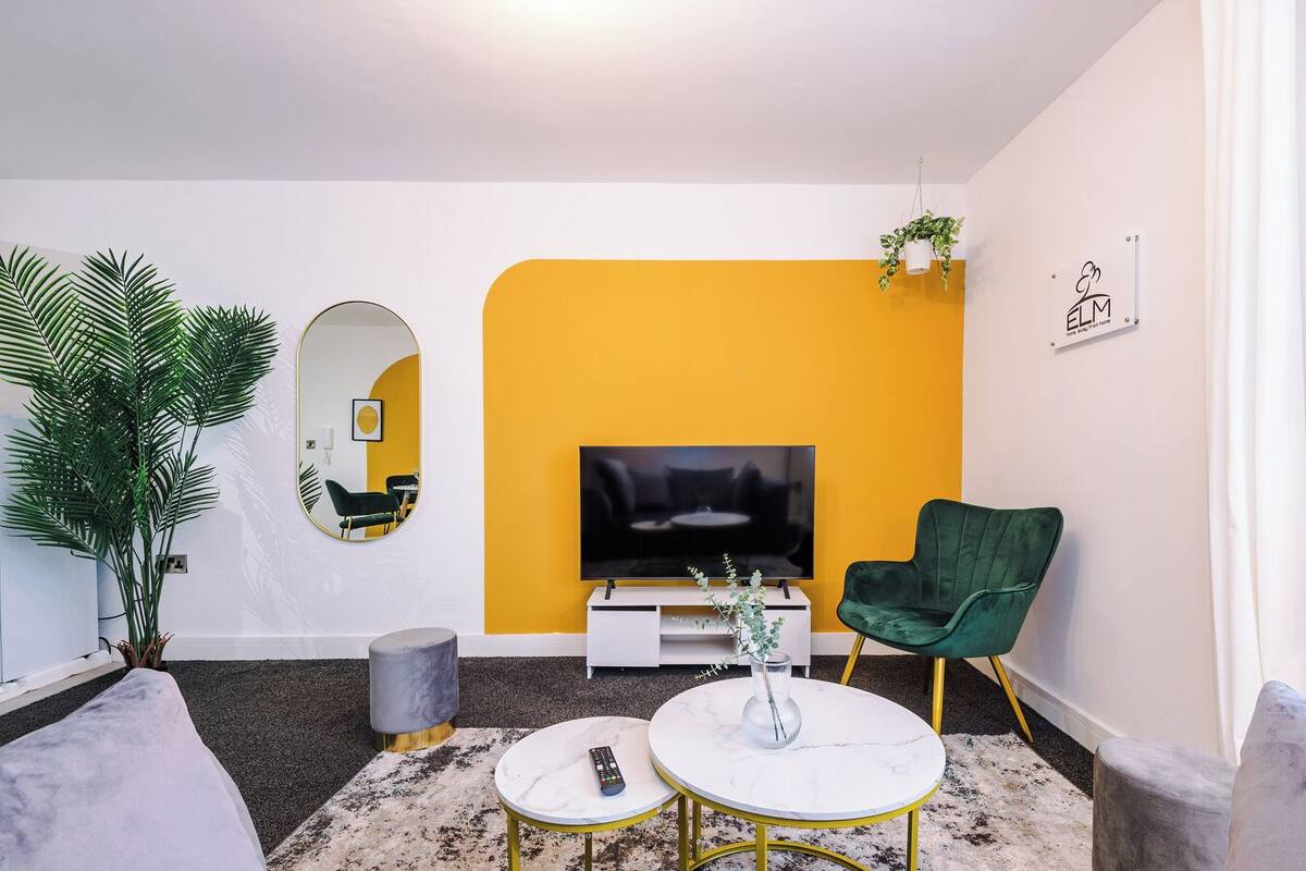 Un salon moderne avec un mur d’accent jaune, un téléviseur à écran plat, un fauteuil vert et une table basse ronde.