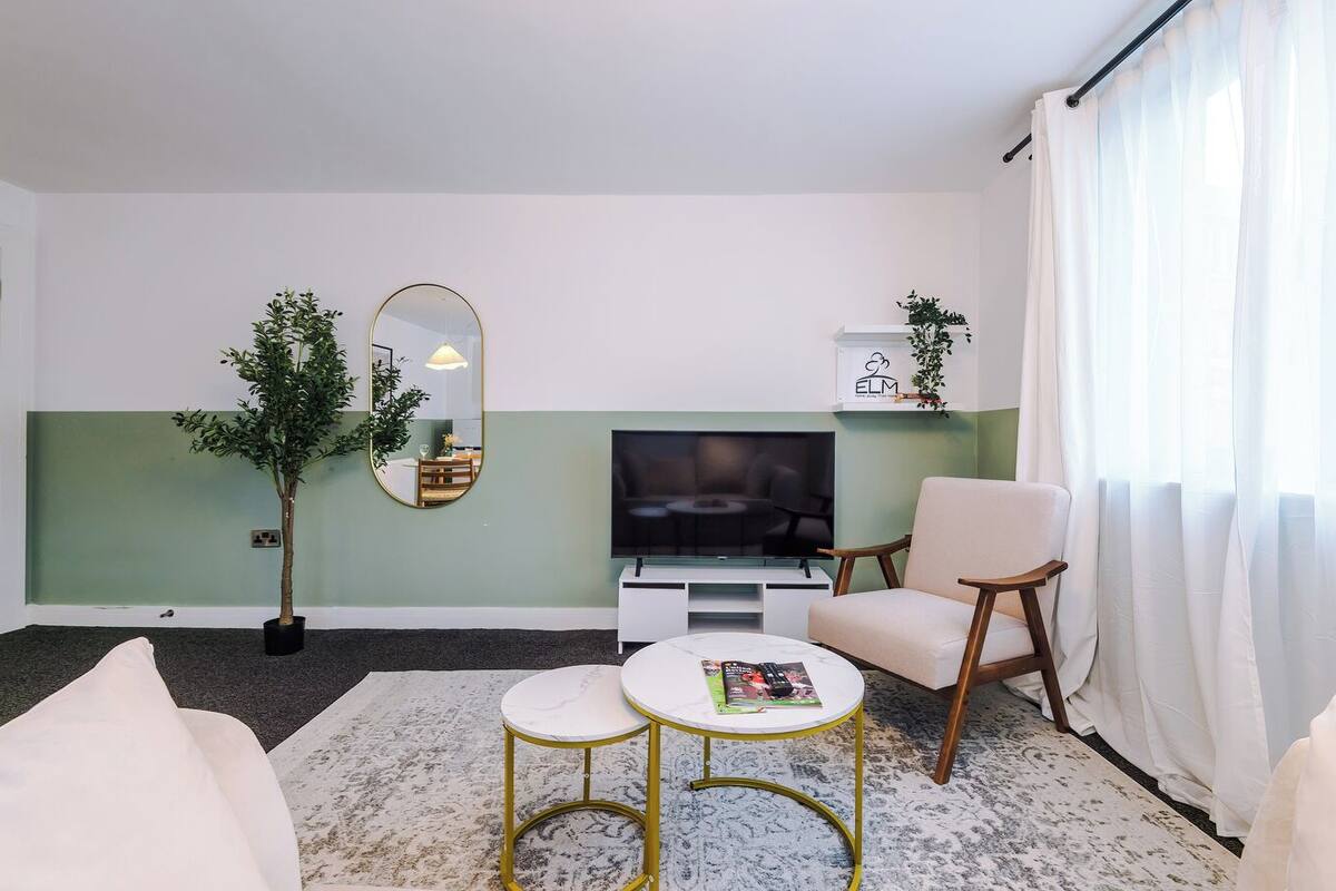 Un salon moderne avec un téléviseur à écran plat, un canapé, un fauteuil, une table basse, un miroir et une plante en pot.