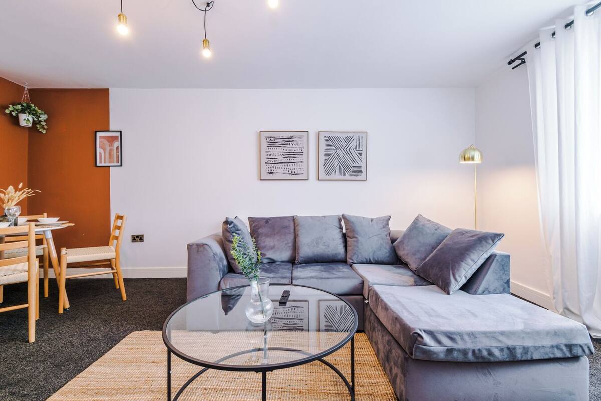 Un salon moderne avec un canapé d’angle gris, une table basse en verre et un coin repas comprenant une table et des chaises en bois.