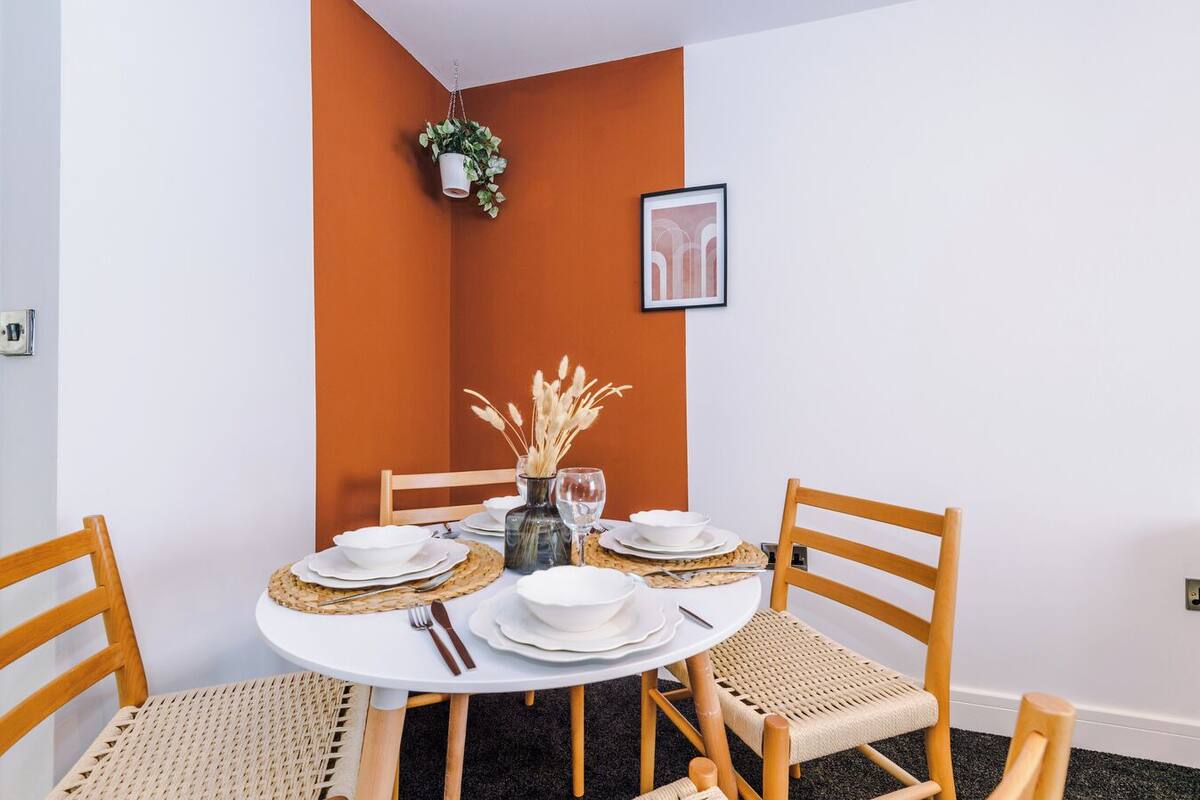 Un coin repas avec une table ronde dressée pour quatre personnes, entourée de chaises en bois dont les assises sont en osier tressé. Une plante en pot est suspendue au plafond, et un tableau encadré est accroché au mur.