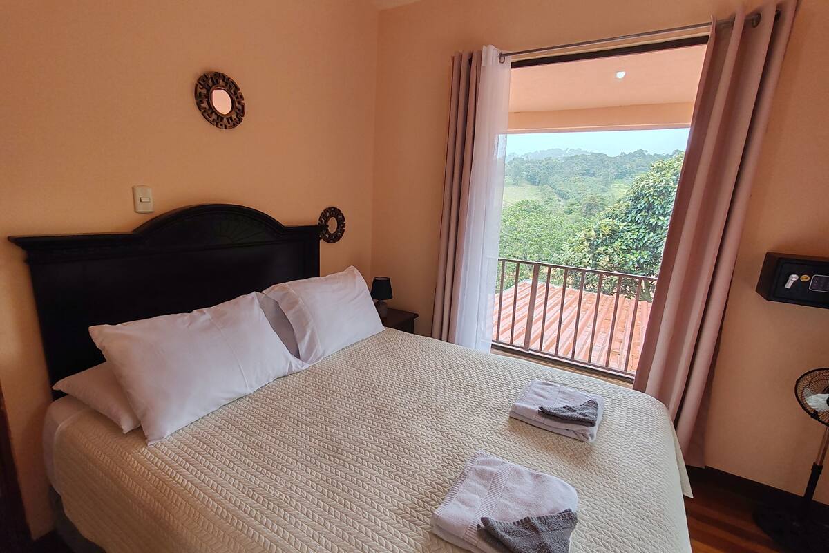 A hotel room with a bed, a view of the outdoors, a nightstand, and a wall-mounted light switch.