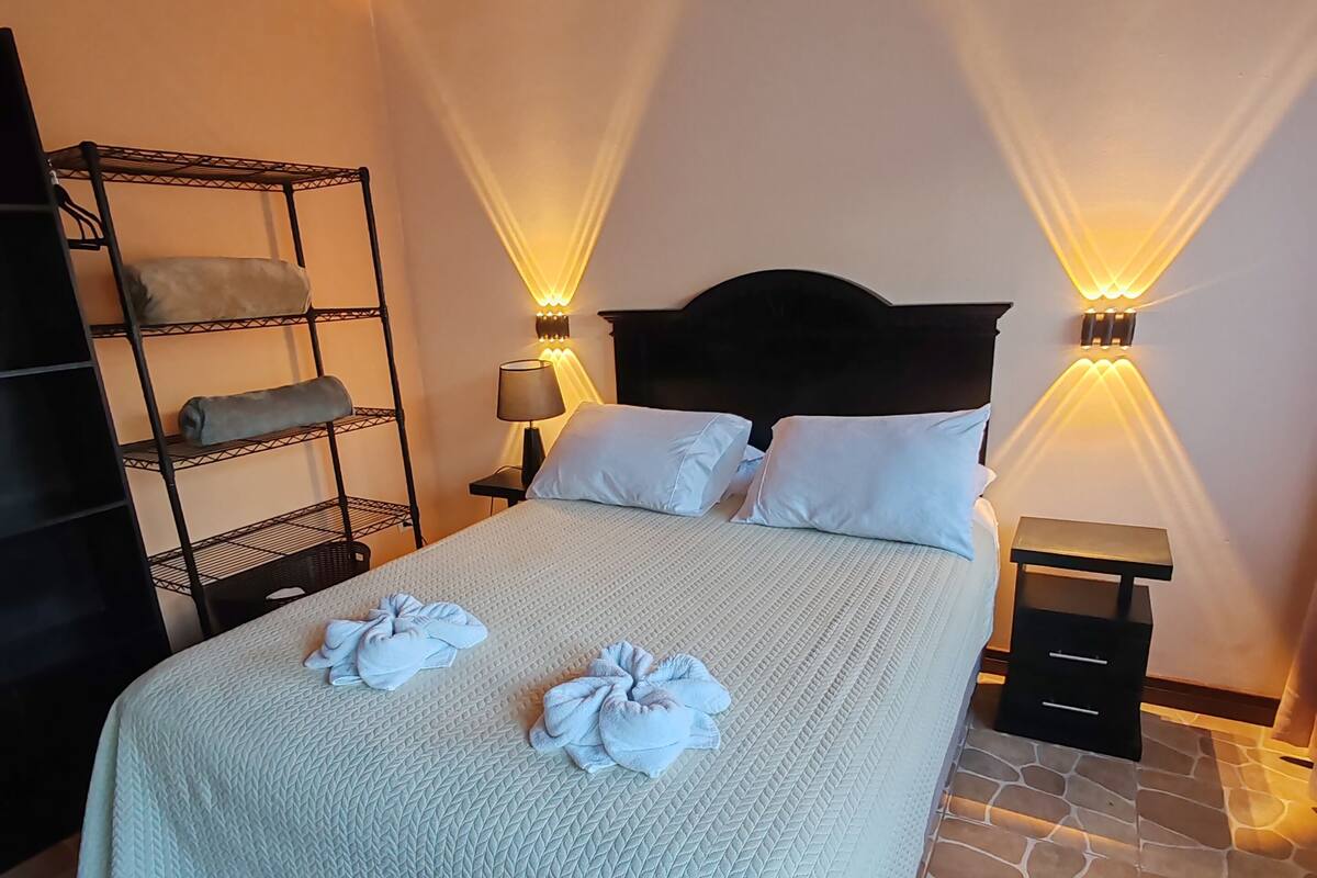 A neatly made bed with a quilted bedspread, two bedside lamps, a nightstand, and a metal shelf unit with folded towels.