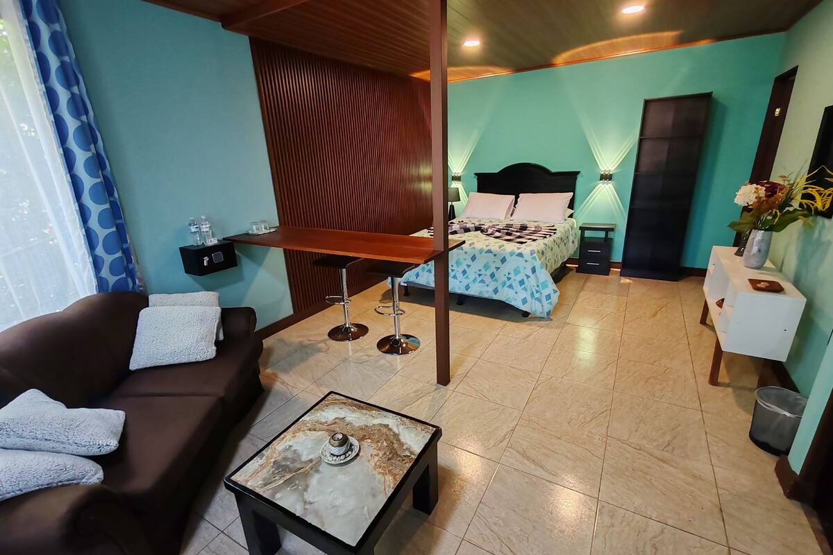 A hotel room with a sofa, a small table, a bed with a patterned bedspread, a nightstand, a wall-mounted shelf with toiletries, and a window with curtains.