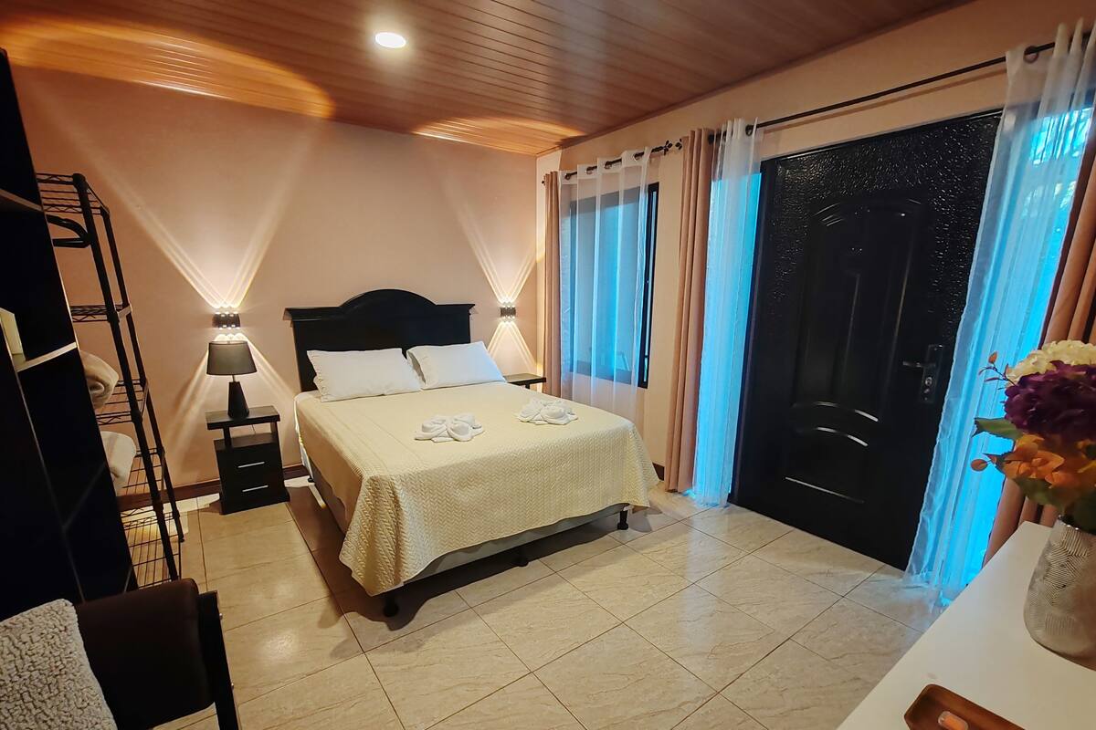 A hotel room with a bed, a desk, a chair, a nightstand, a lamp, a window with curtains, and a door.