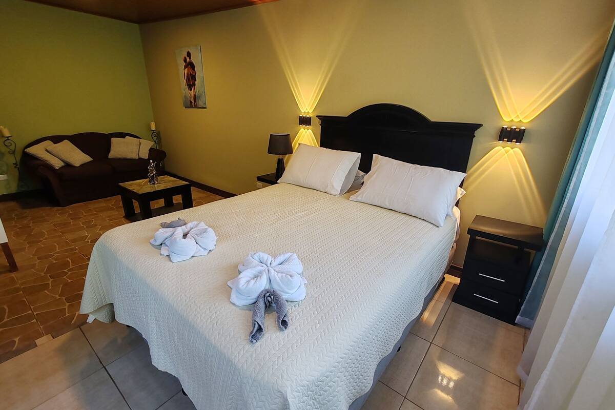 A neatly made bed with towels, a dark headboard, bedside lamps, a small table, and a sofa in a room with tiled flooring and a green wall.