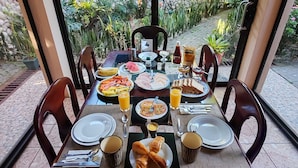 Mesa de jantar posta com pratos, talheres e diversos pratos, rodeada por cadeiras, com vista para um jardim através de uma divisória de vidro.