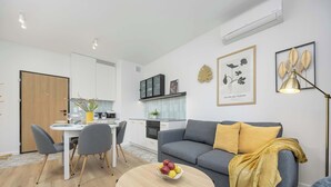 Uma sala de estar moderna com um sofá cinzento, uma mesa de centro redonda de madeira com frutas, uma área de jantar com uma mesa branca e cadeiras cinzentas, e uma cozinha ao fundo.
