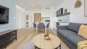 Uma sala de estar moderna com um sofá cinza, uma mesa de centro redonda de madeira com frutas, uma televisão na parede e uma cozinha ao fundo.