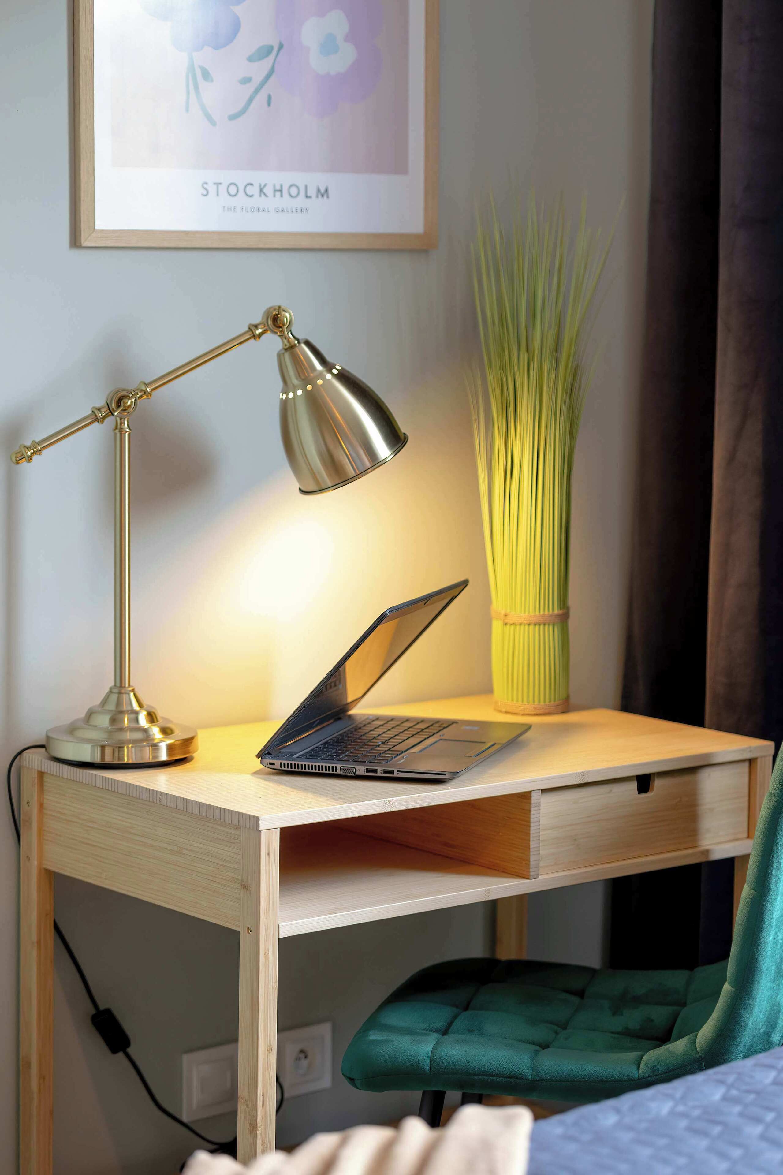 A wooden desk with a laptop, a desk lamp, and a tall vase with green plants.