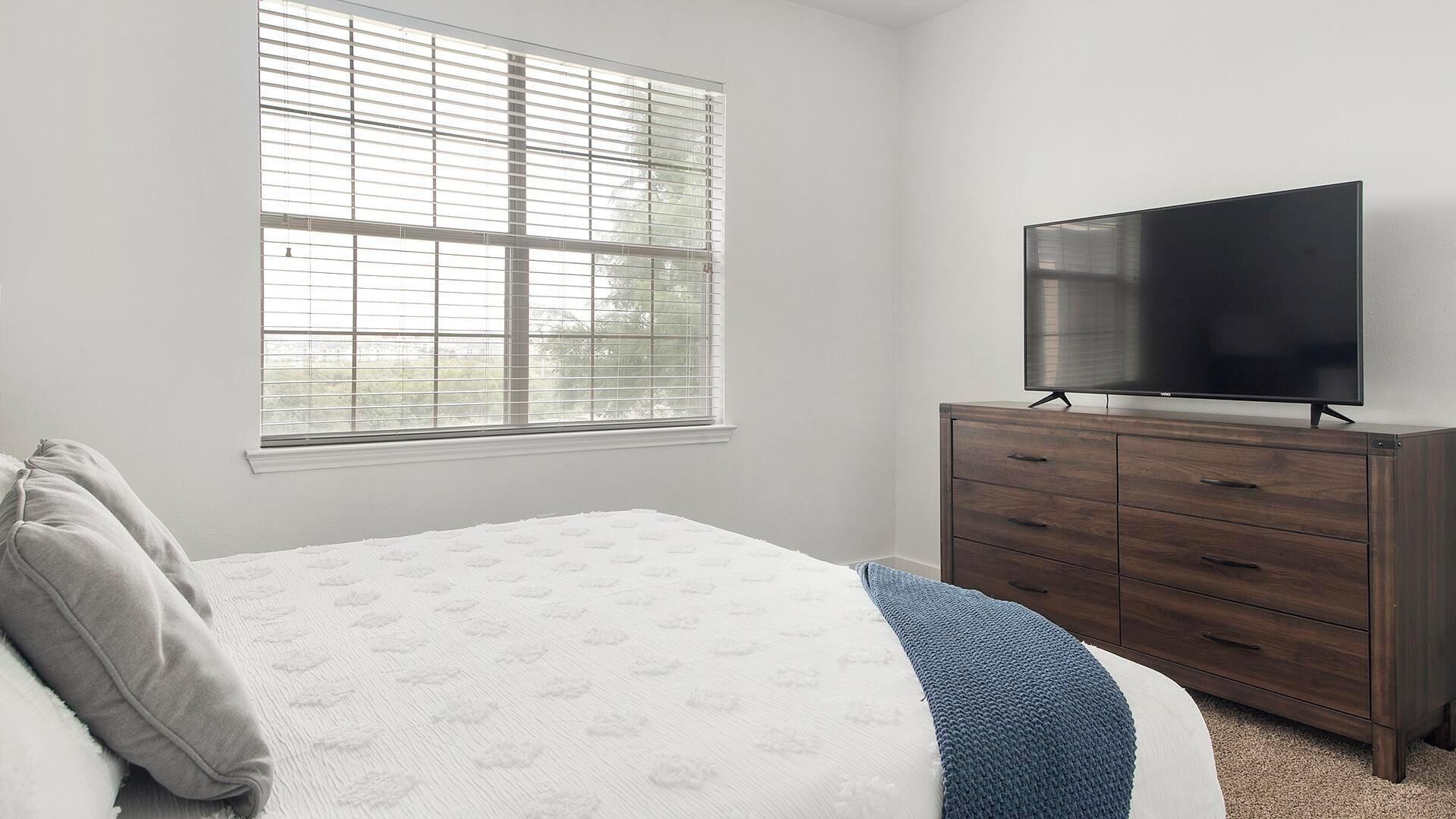 A bedroom with a bed, a dresser, a TV, and a window with blinds.