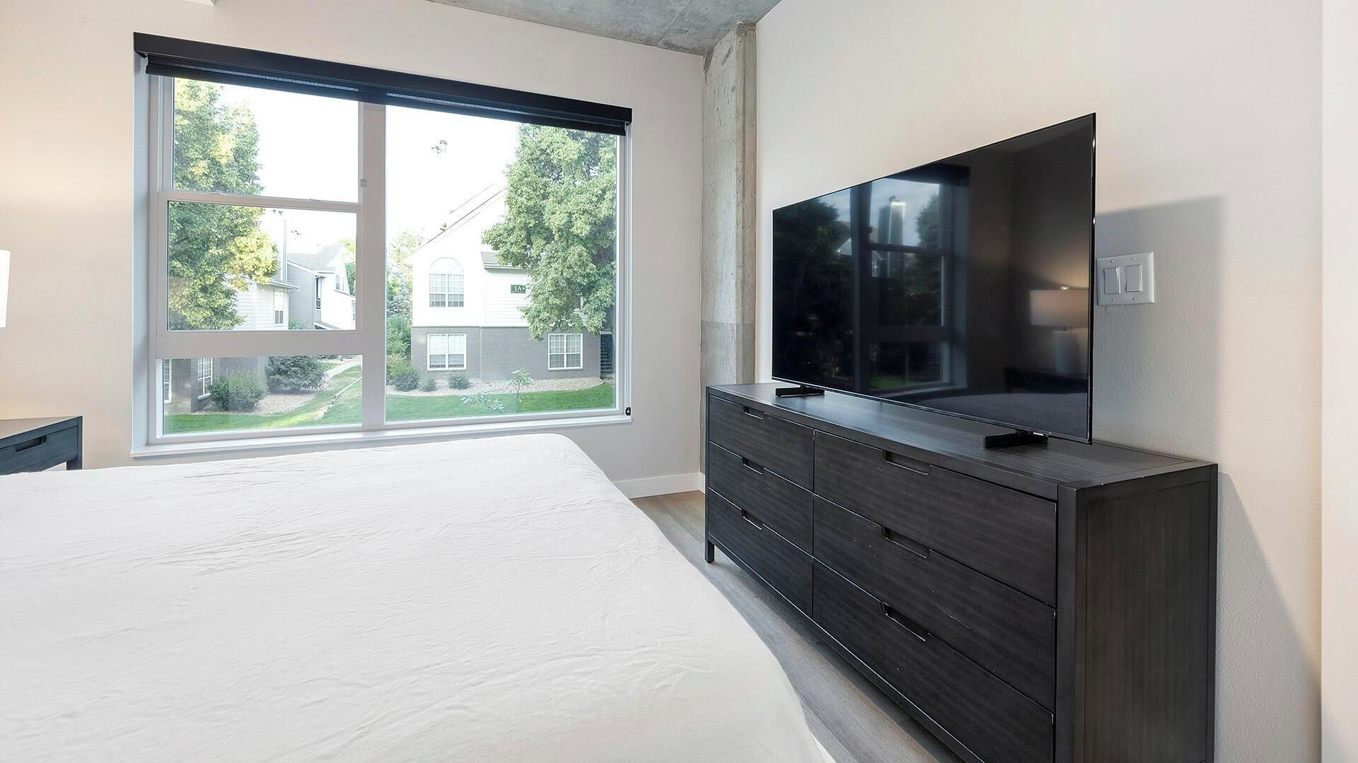 Ett sovrum med en stor säng, en mörk byrå, en platt-TV och ett fönster med utsikt över ett hus och träd.