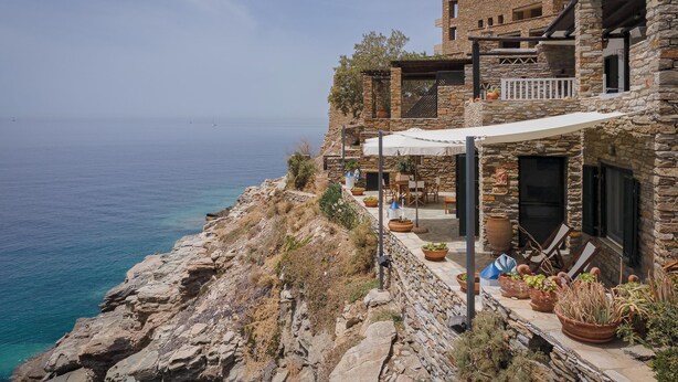 Un bâtiment en pierre doté d’une terrasse offrant une vue sur la mer, avec des espaces de repos en plein air et des plantes en pot.