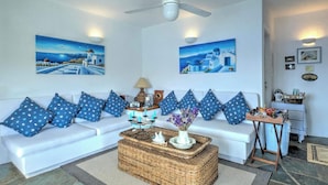 Un salon avec un canapé blanc, des coussins à motifs bleus, une table basse en osier agrémentée d’objets décoratifs, et deux tableaux représentant des paysages côtiers, accrochés au mur.