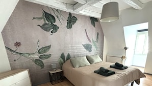 A bedroom with a bed, a nightstand, a dresser, a hanging light fixture, and a window. - Charming apartment with character (Nevers)