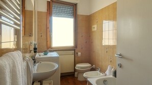 Et badeværelse med vask, toilet og badekar. Der er et vindue med persienne, og gulvet er fliset.