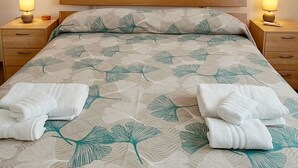 En pænt redt seng med et sengetæppe med bladmotiv, to sengeborde med lamper og to sengeborde med skuffer.