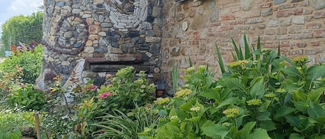 Un mur de pierre orné d’un motif circulaire, une fenêtre avec des volets et diverses plantes dans un jardin.
