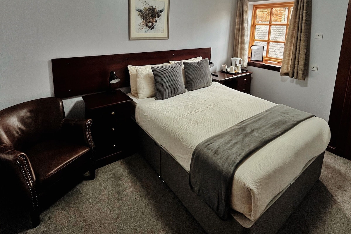 A bedroom with a large bed, a wooden headboard, a brown leather sofa, a bedside table with a lamp, and a framed picture on the wall.