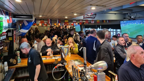 Un bar affollato con un grande schermo che mostra un evento sportivo, una fontana di birra e diverse bandiere.
