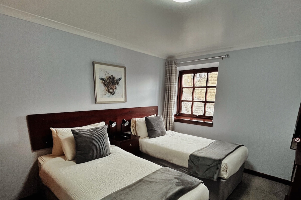 A hotel room with two beds, a window with curtains, a framed picture on the wall, and a ceiling light.