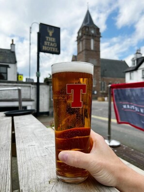 A hand holding a glass of beer with a 'T' logo, in front of a building with a clock tower and a sign that reads 'The Stad Hotel'.