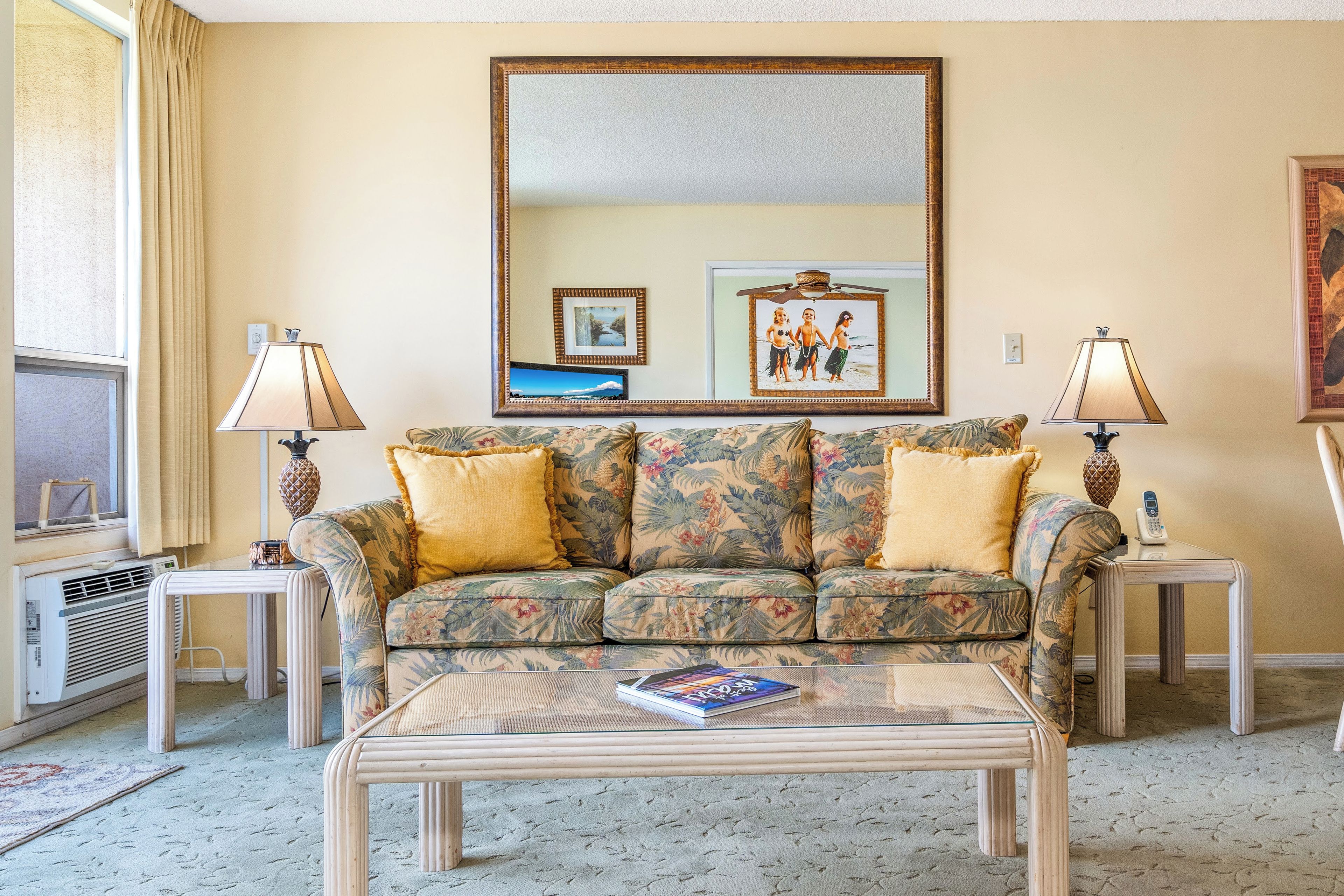 A living room with a floral sofa, two lamps, a coffee table with a book, a large mirror, and a framed picture on the wall.