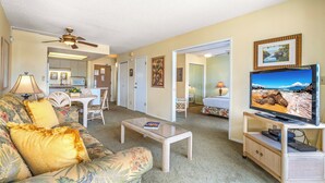 A living room with a sofa, coffee table, television, and a ceiling fan. There is a kitchen area in the background.