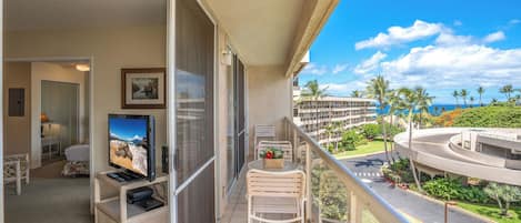A balcony with a TV, seating, and a view of a landscaped area with palm trees and a building.