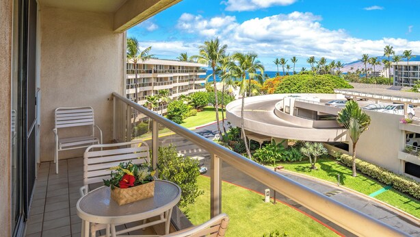 A balcony with a table and chairs, overlooking a landscaped area with palm trees and a view of a building complex.