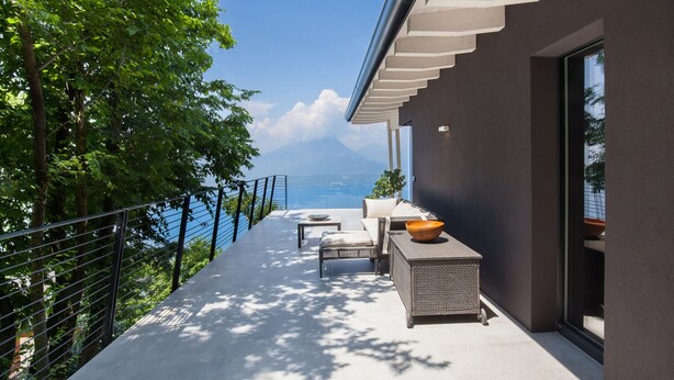 A modern outdoor balcony with a view of the sea and mountains, furnished with a sofa, table, and chairs.