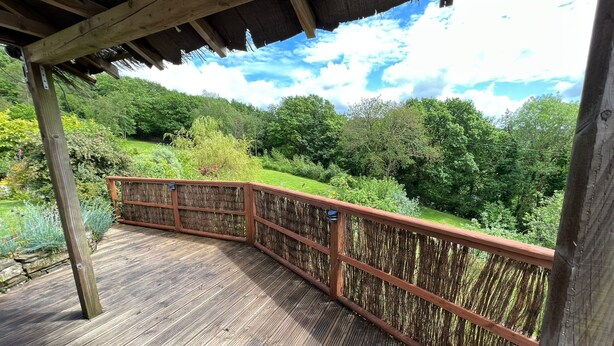 A wooden deck with a thatched roof, surrounded by lush greenery and a clear sky.