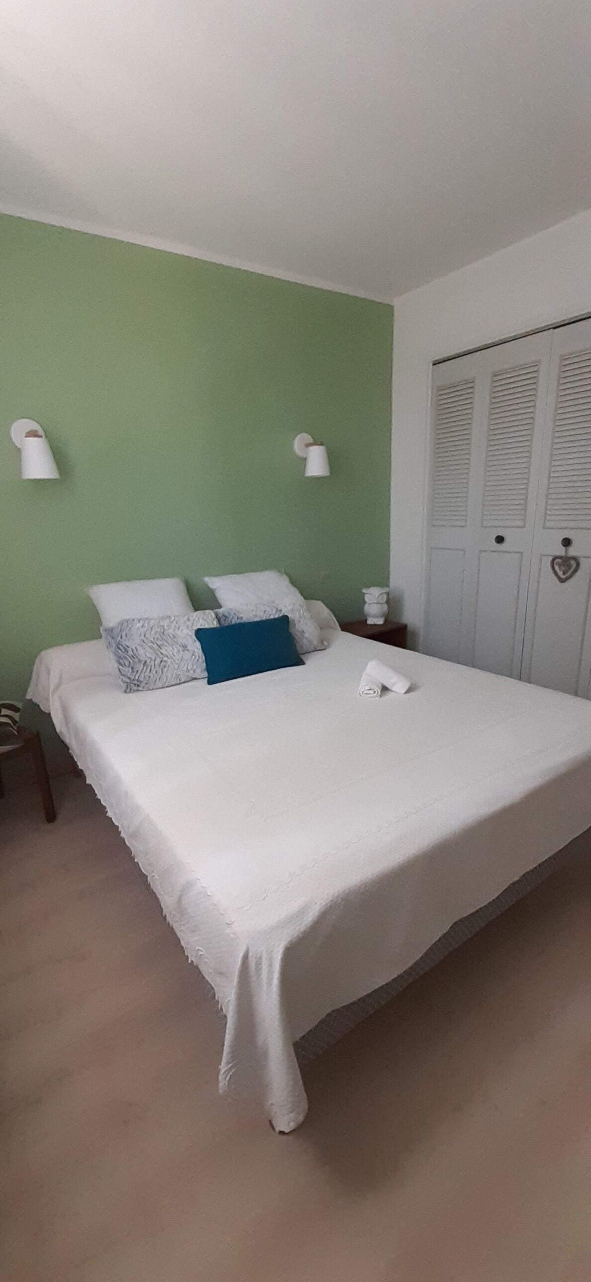 Un lit bien fait, avec une literie blanche et un oreiller bleu, une table de chevet avec une lampe blanche et un mur vert.