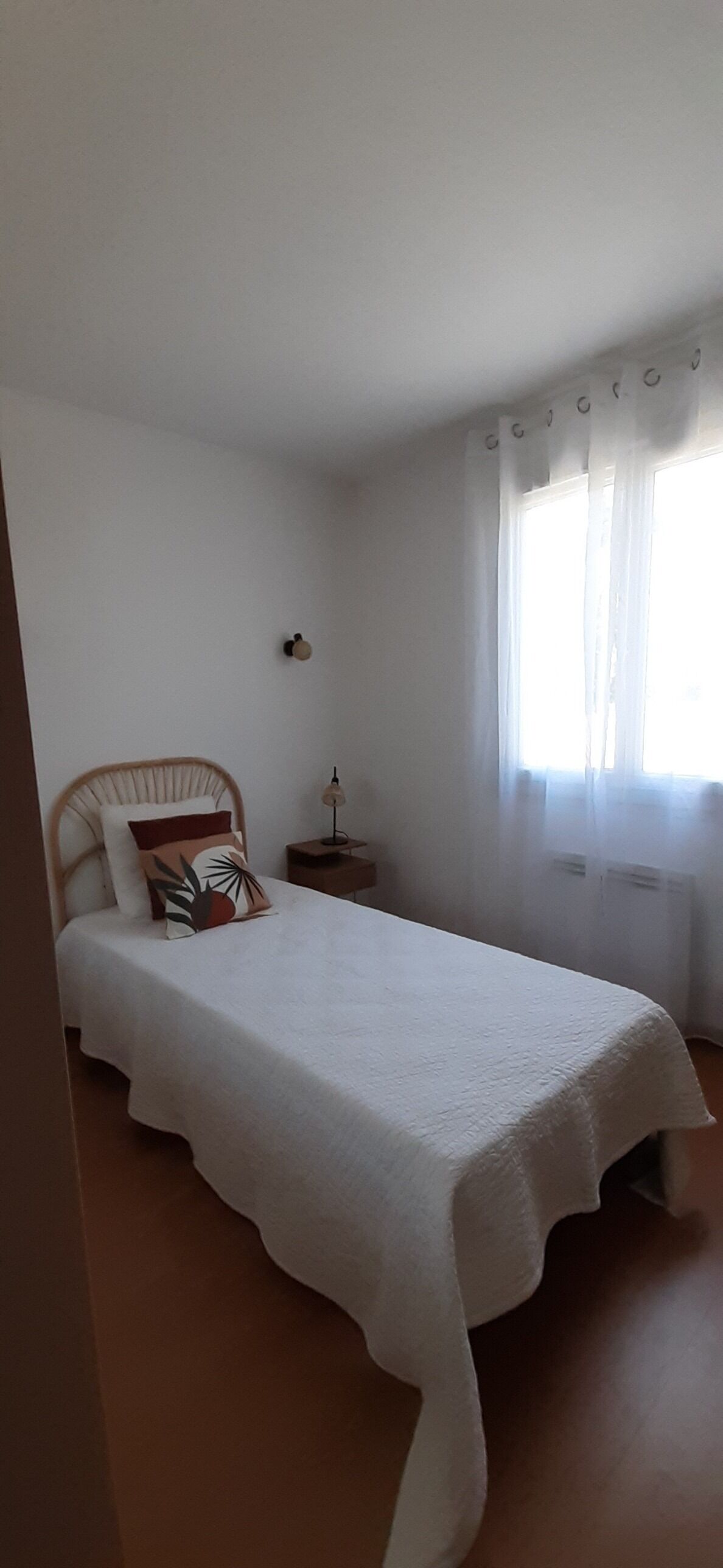 A bedroom with a single bed, a nightstand, a lamp, and a window with sheer curtains.